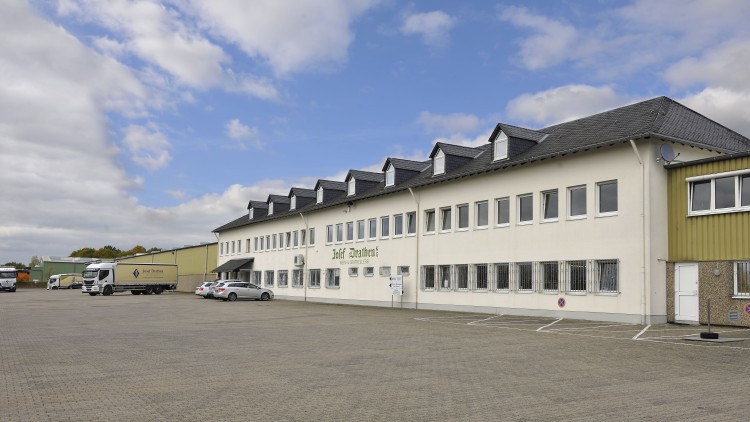 Ein zweistöckiges Gebäude mit weißen Wänden, Parkplatz im Vordergrund und einem Lkw mit der Aufschrift „Josef Drathen“.