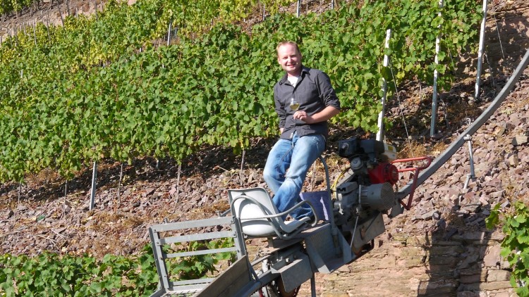 Person sitzt mit einem Glas Weißwein auf einer motorisierten Schienenbahn in steilem Weinberg mit grünen Reben.