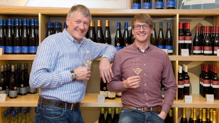 Zwei Personen stehen vor Weinregalen und halten jeweils ein Glas Weißwein in der Hand, eine Wandkarte ist im Hintergrund.