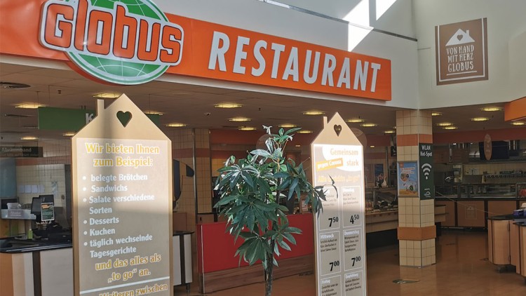 Eingangsbereich des GLOBUS Restaurants mit orangem Schriftzug, Speisetafeln, Pflanzen und Hinweisen zu „to go“-Angeboten.