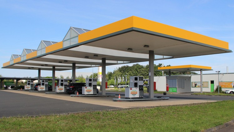 GLOBUS Tankstelle mit gelbem Dach, mehreren Zapfsäulen und geparktem Auto auf asphaltierter Fläche vor einer grünen Umgebung.