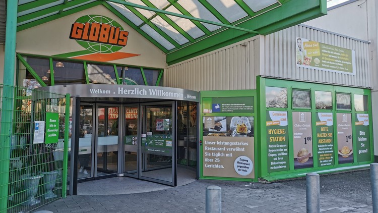 Eingangsbereich eines GLOBUS Marktes mit grünem Rahmen, Drehkreuz und Werbeschildern mit Hinweisen zu Restaurant und Hygiene.