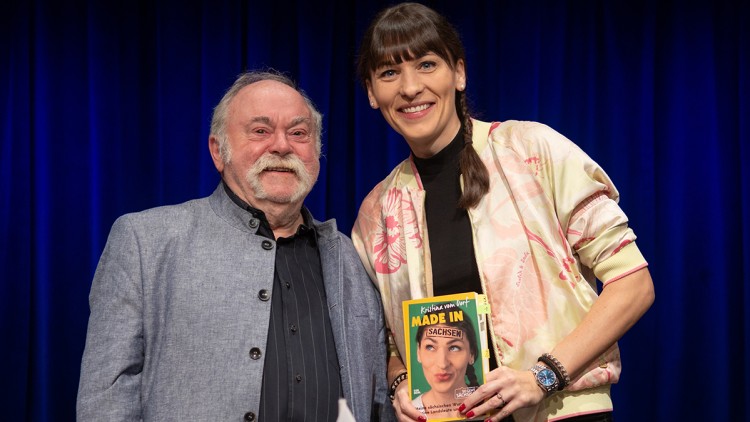 Zwei Personen vor einem dunkelblauen Vorhang, eine hält das Buch „Krisha von der Loff, MADE IN“ mit gelb-grünem Cover.