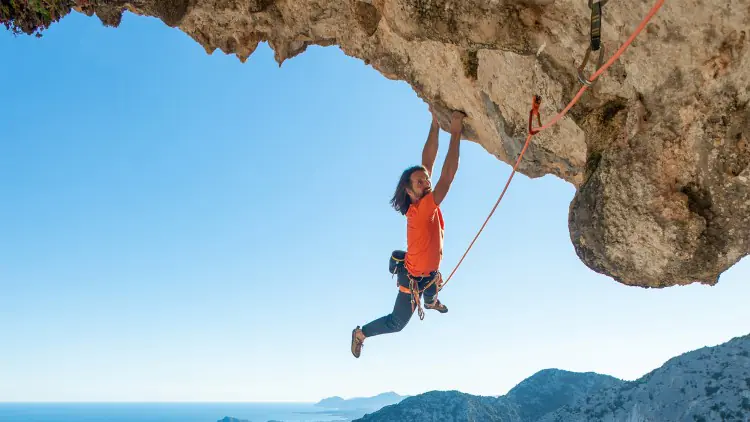 Person mit orangefarbenem Shirt klettert an einer Felswand mit Kletterseil, im Hintergrund Berge und Meer unter blauem Himmel.