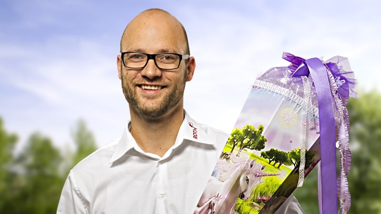 Eine Person hält eine bunte Schultüte mit Einhornmotiv, lila Schleife und Blumen vor einer grünen Wiesenlandschaft.