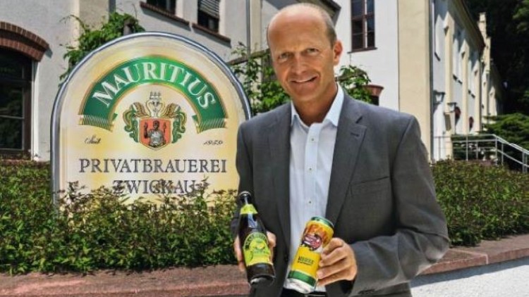 Eine Person in grauem Anzug hält eine Bierflasche und eine Dose vor einem Schild der Mauritius Privatbrauerei Zwickau.