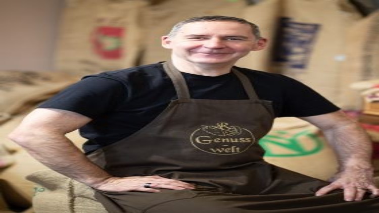 Eine Person sitzt vor Kaffeesäcken und trägt eine braune Schürze mit der goldenen Aufschrift „Genusswelt“.