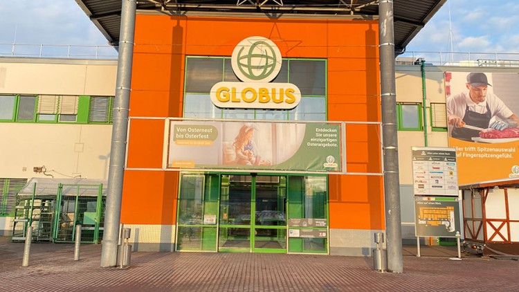 Eingangsbereich eines GLOBUS-Marktes mit orangener Fassade, grünem Logo, Glasdrehtür und Werbebannern an der Front.