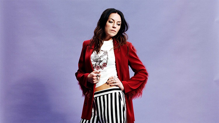 Person in rotem Blazer mit Fransen, weißem bedrucktem T-Shirt und schwarz-weiß gestreifter Hose vor lila Hintergrund.