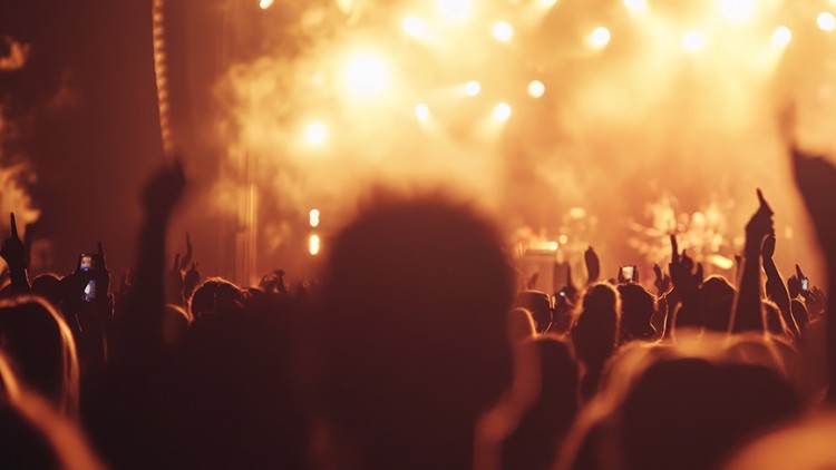 Menschenmenge bei einem Konzert mit hochgereckten Händen vor einer beleuchteten Bühne mit warmen, gelblichen Strahlern.