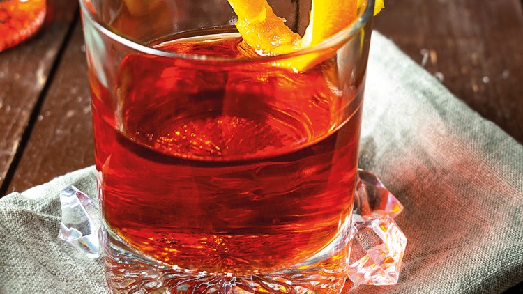 Ein quadratisches Glas mit rotem Getränk und orangefarbenen Zesten steht auf einem grauen Stofftuch auf Holztisch.