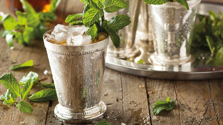 Silberbecher mit Eiswürfeln, Getränk und frischer Minze, auf einem Holztisch mit Minzblättern und einem Tablett im Hintergrund.