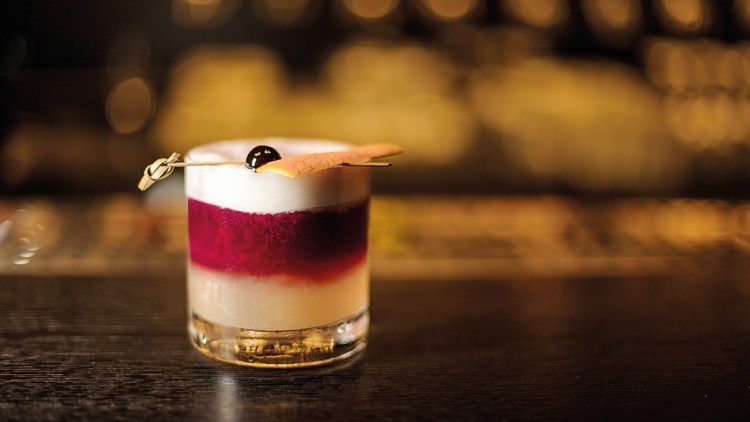 Ein Cocktailglas mit weißem Schaum, roter Schicht und Deko aus Kirsche und Keks auf dunklem Tresen, Hintergrund unscharf.