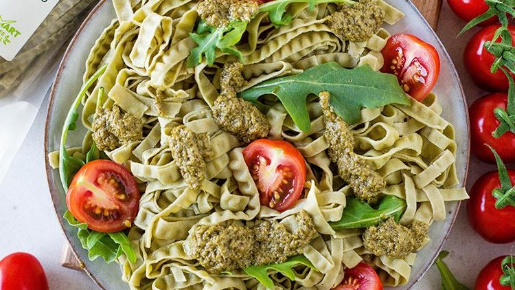Grüne Nudeln mit Pesto, halbierten Cherrytomaten und Rucola auf einem Teller, daneben Kirschtomaten, Cashewnüsse und Verpackung.