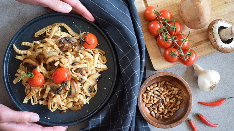 Ein schwarzer Teller mit Pasta, gebratenen Pilzen und Tomaten, daneben Zutaten wie Knoblauch, Pinienkerne und Chilis.