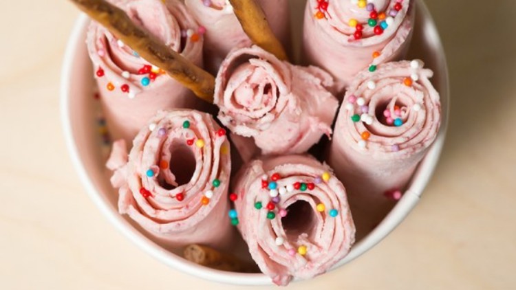 Gerolltes rosa Eis mit bunten Zuckerstreuseln und zwei Salzstangen in einem weißen Pappbecher, daneben ein Holzlöffel.