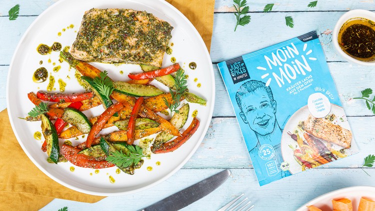 Ein Teller mit gebratenem Lachs, Gemüse und Kräutern neben einer Gewürzpackung mit der Aufschrift „Moin Moin“ auf hellem Holztisch.