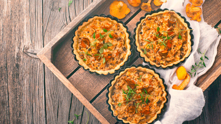Drei gebackene Mini-Quiches mit Kräuterdekoration und Pfifferlingen in einer Holzschale auf rustikalem Untergrund.