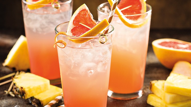 Drei Gläser mit rosa Getränk, Eiswürfeln und Scheiben von Grapefruit und Ananas, umgeben von Ananas- und Grapefruitstücken.