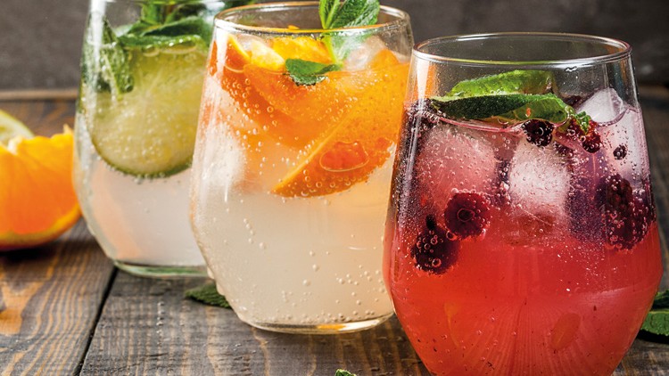 Drei Gläser mit sprudelnden Getränken und Eiswürfeln, jeweils mit Minze, Früchten wie Beeren, Orangen und Gurkenscheiben dekoriert.