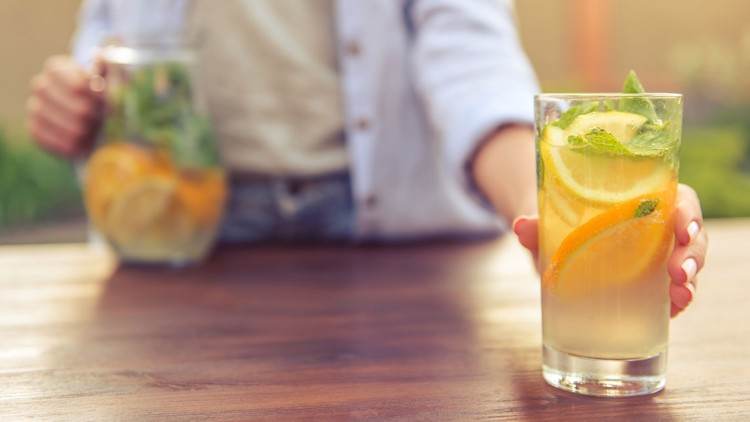 Ein Glas mit Getränk aus Wasser, Zitronen-, Orangen- und Limettenscheiben sowie Minze wird auf einem Holztisch gereicht.