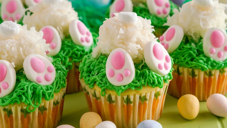 Cupcakes mit grünem Zuckerguss, Kokosraspeln, rosa Hasenpfoten-Dekor und kleinen bunten Schoko-Ostereiern auf grünem Tisch.
