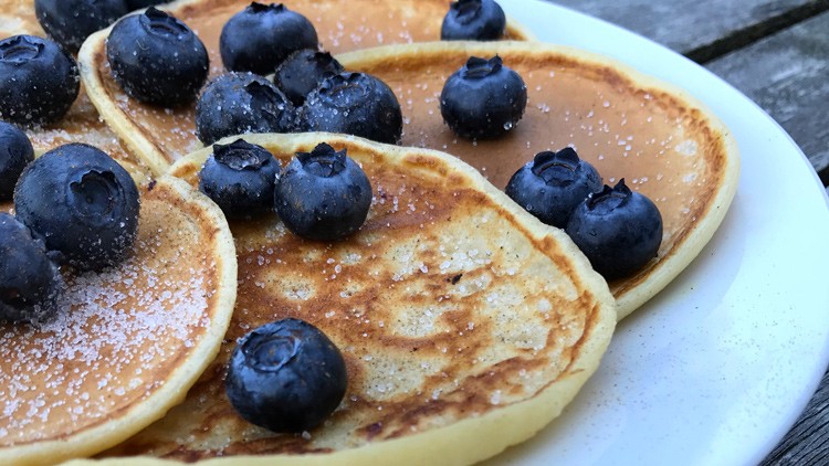 Fünf goldbraune Pancakes mit frischen Blaubeeren und Zuckerkristallen auf einem weißen Teller.