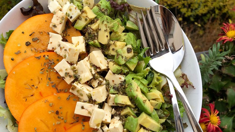 Salatschüssel mit grünen Blättern, Persimonenscheiben, gewürfeltem Käse, Avocado, Kräutern und Besteck auf dem Rand.