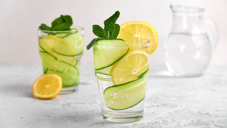 Zwei Gläser Wasser mit Gurken-, Zitronenscheiben und Minzblättern stehen auf hellem Untergrund, daneben eine Karaffe Wasser.